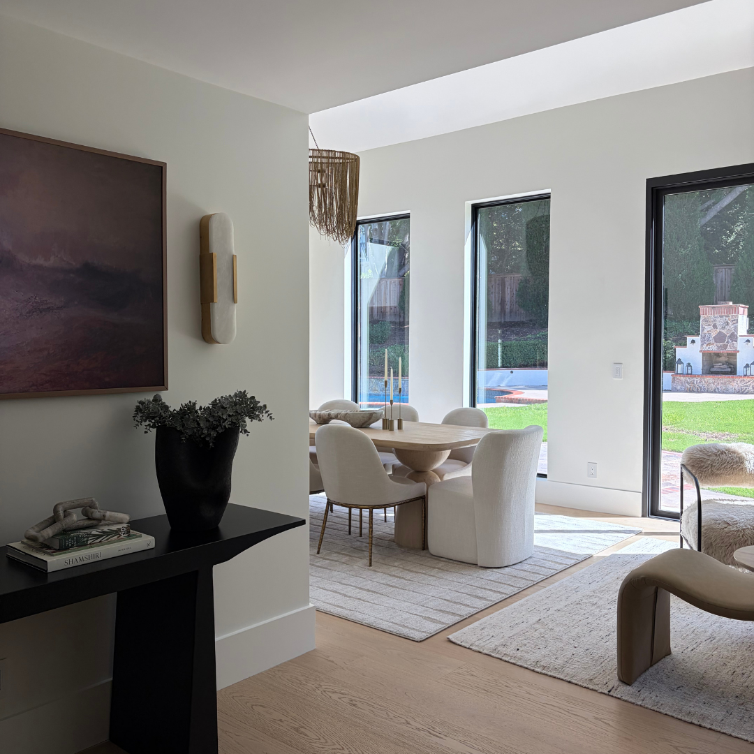 A modern interior space with light wood flooring, white walls, and large windows. In the foreground, a black console table holds a dark vase with greenery and a framed artwork.