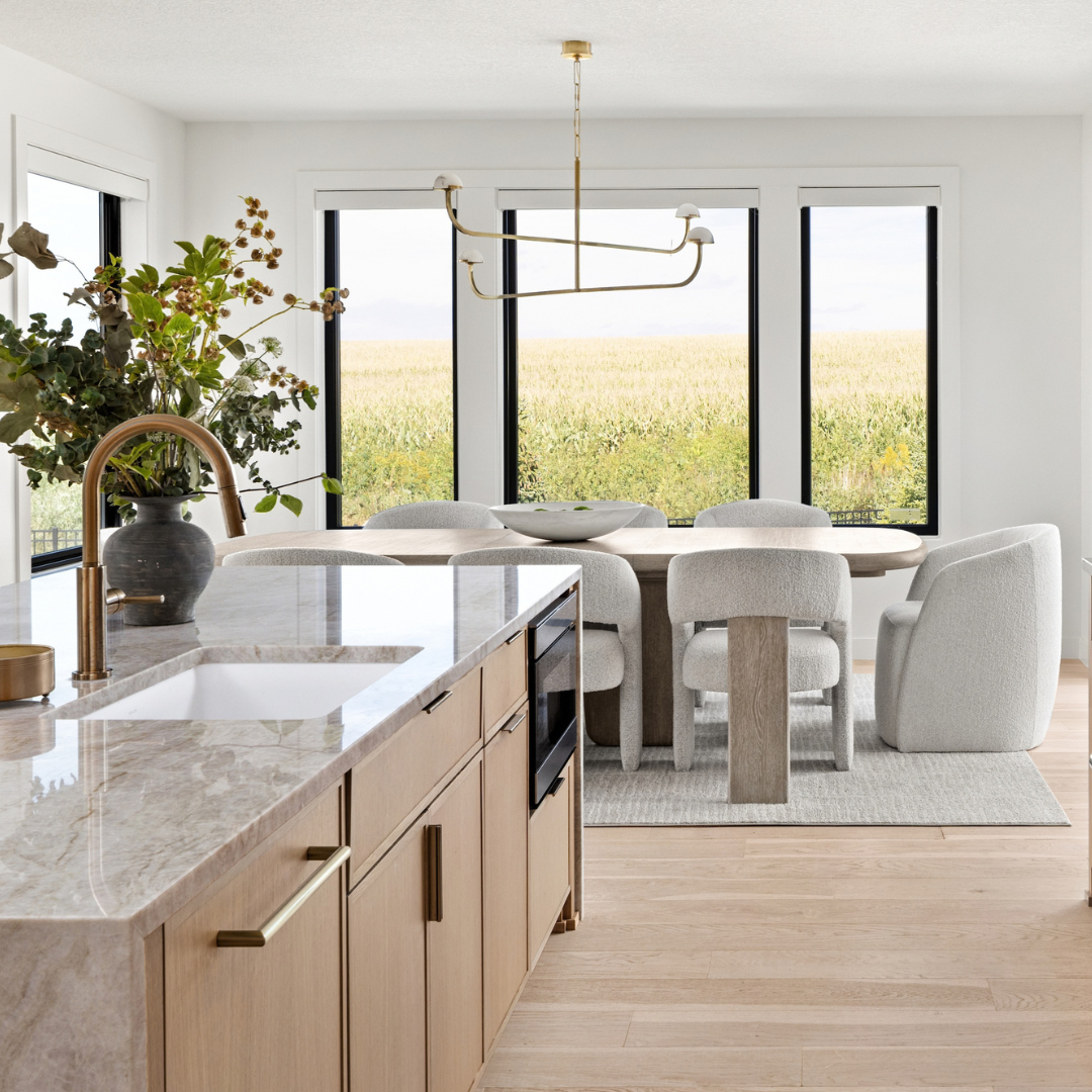 A modern kitchen island with light wood cabinets, a marble countertop, and a curved brass faucet, shown in a bright, contemporary dining room with large windows overlooking a field.