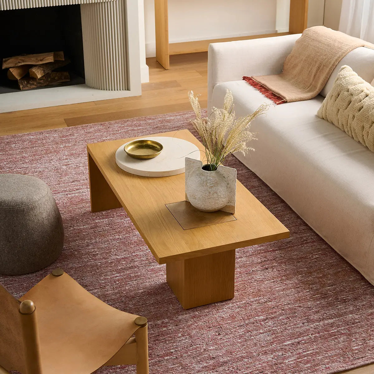 A modern light wood coffee table in a styled living room setting, placed on a textured pinkish-red area rug next to a white sofa and in front of a fireplace.
