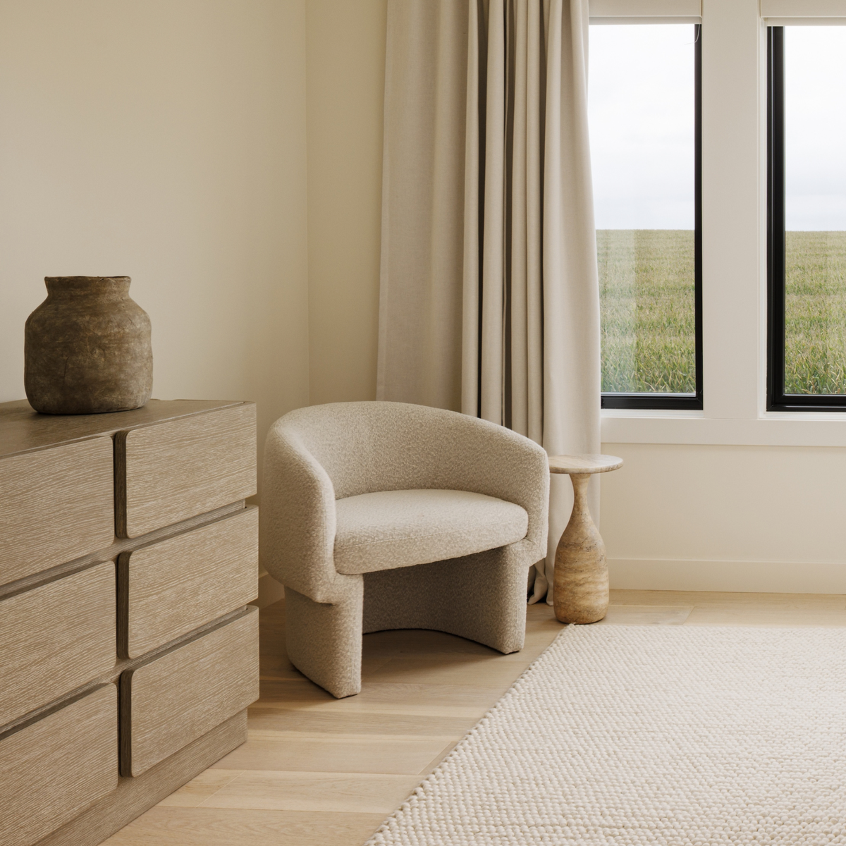 A modern upholstered accent chair in a textured beige bouclé fabric, positioned in a minimalist room next to a light wood dresser and a concrete side table.