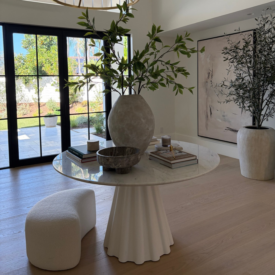 A modern round marble-top dining table with a sculptural fluted pedestal base, styled in a bright contemporary room with a large textured gray vase, books, and a small white pouf. Large glass doors in the background reveal a garden, and a large abstract print hangs on the wall.
