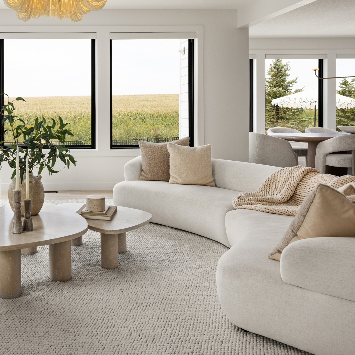 A modern curved white sectional sofa in a bright, contemporary living room with beige throw pillows, a textured rug, and large windows overlooking a grassy field.