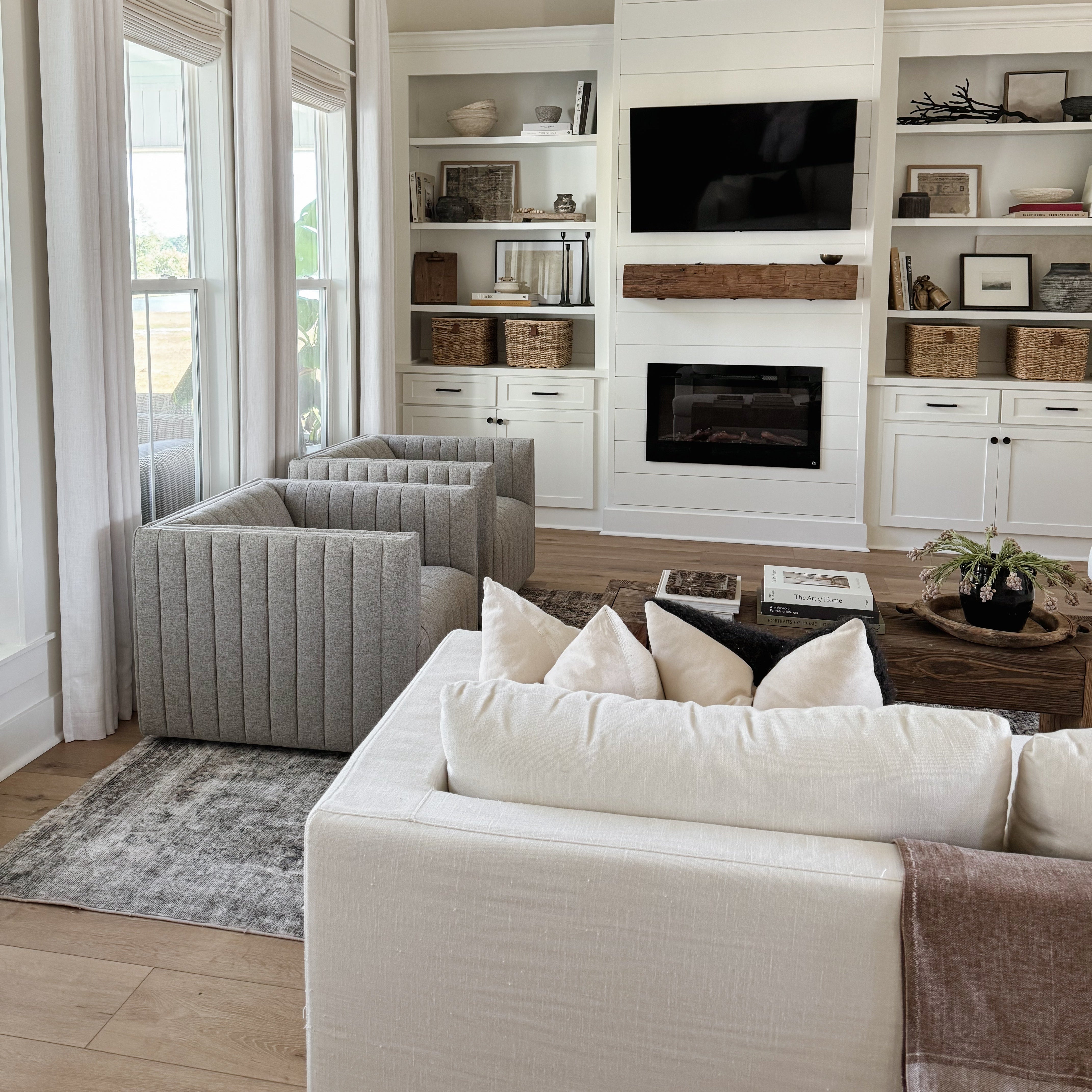 A bright, modern living room featuring a white shiplap wall, built-in shelves with decorative baskets and framed art, a black TV and fireplace, gray tufted armchairs, and a white sofa in the foreground.
