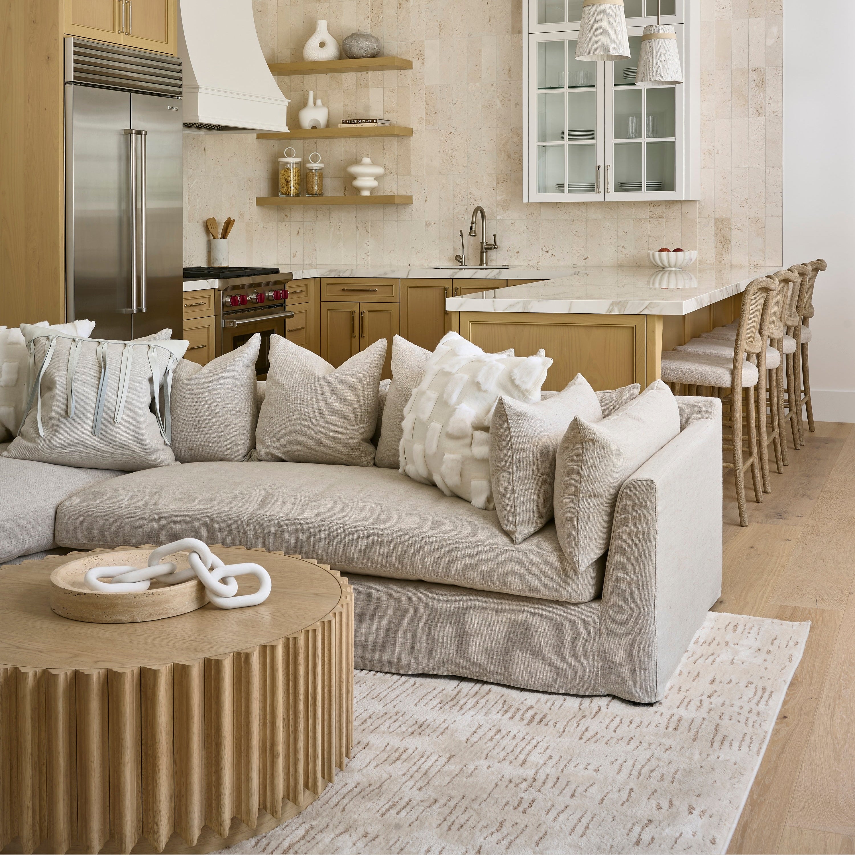 A modern living room featuring a large light beige sectional sofa with textured throw pillows, a round ribbed wooden coffee table, and a large clear glass chandelier. In the background, a contemporary kitchen with light wood cabinets, a marble island, and a white-framed window is visible.