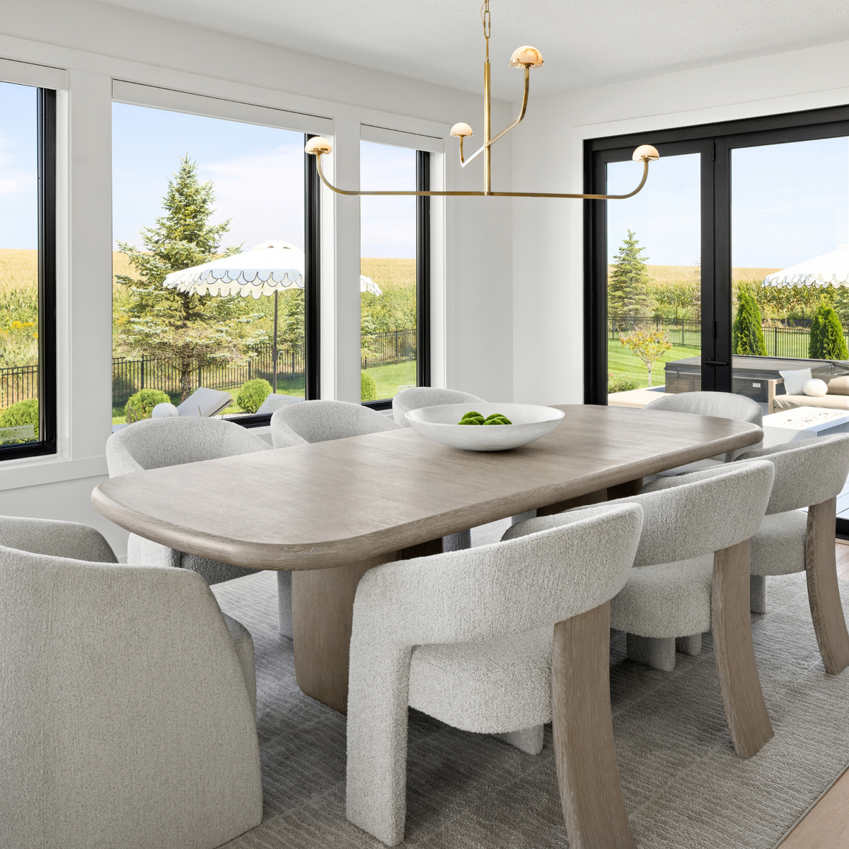 A modern dining room featuring a light oak wood rounded rectangular table surrounded by textured light gray upholstered chairs, with a brass multi-arm chandelier overhead and large windows showing an outdoor landscape.