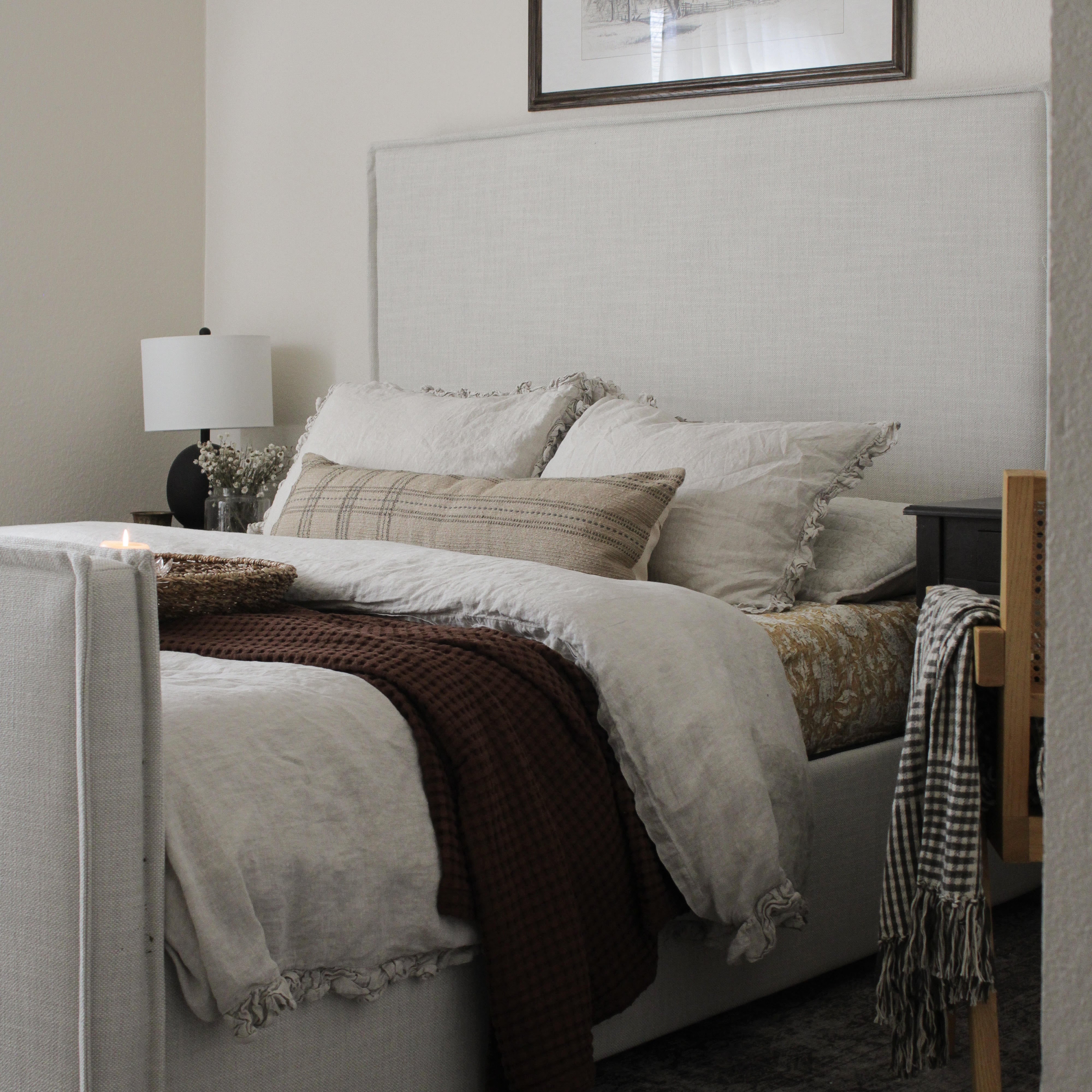 A styled bedroom scene featuring a bed with a tall upholstered headboard, dressed in cream bedding, a dark brown throw blanket, and multiple pillows. A black nightstand with a white lamp sits on the left, and a wooden chair with a black-and-white fringed throw is on the right.