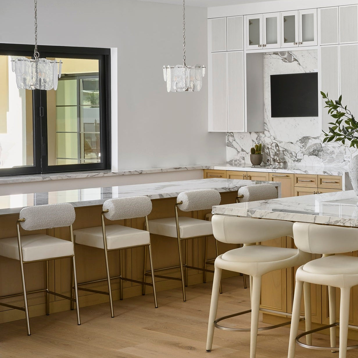 A modern kitchen island with a white marble countertop and backsplash, surrounded by cream bar stools, set in a bright kitchen with light wood cabinets and white walls.