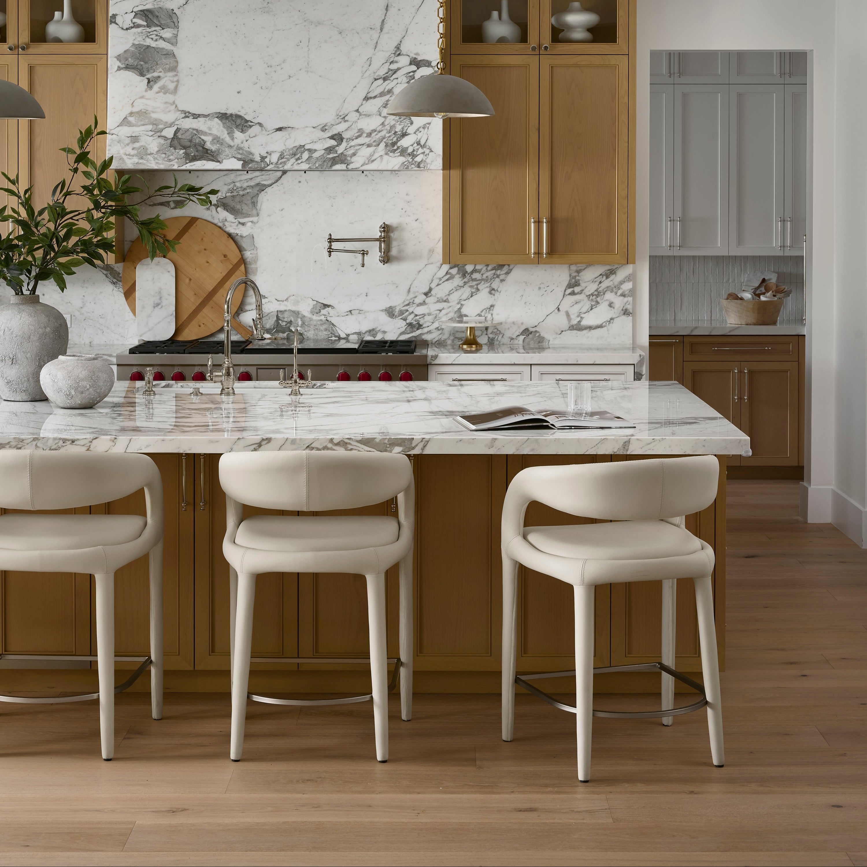 A modern kitchen interior with a large white marble island and matching marble backsplash, light wood cabinetry, pendant lights, and three cream bar stools in the foreground.