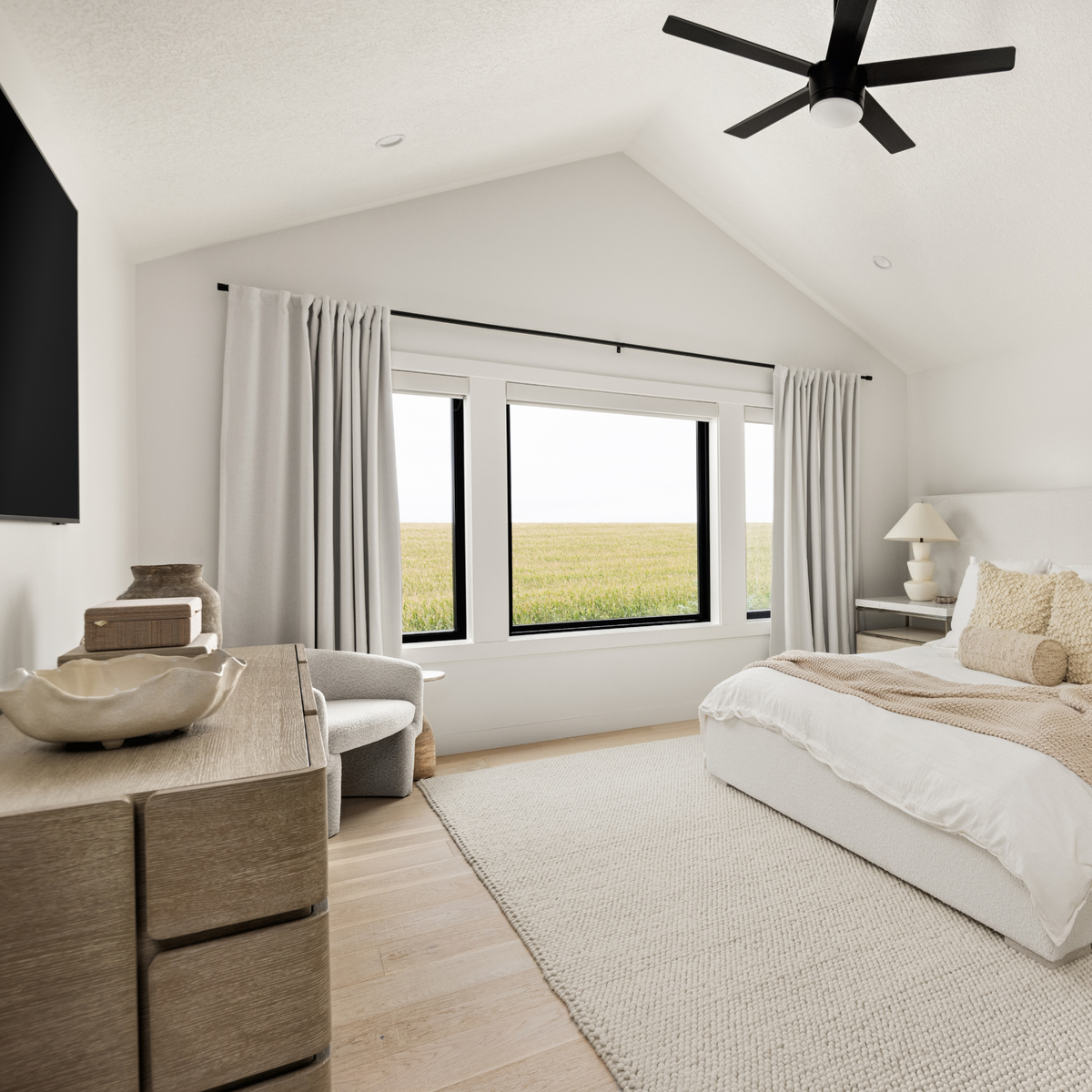 A bright, modern bedroom with a large window overlooking a grassy field, featuring white bedding, a textured beige rug, light wood furniture, and neutral-toned curtains.