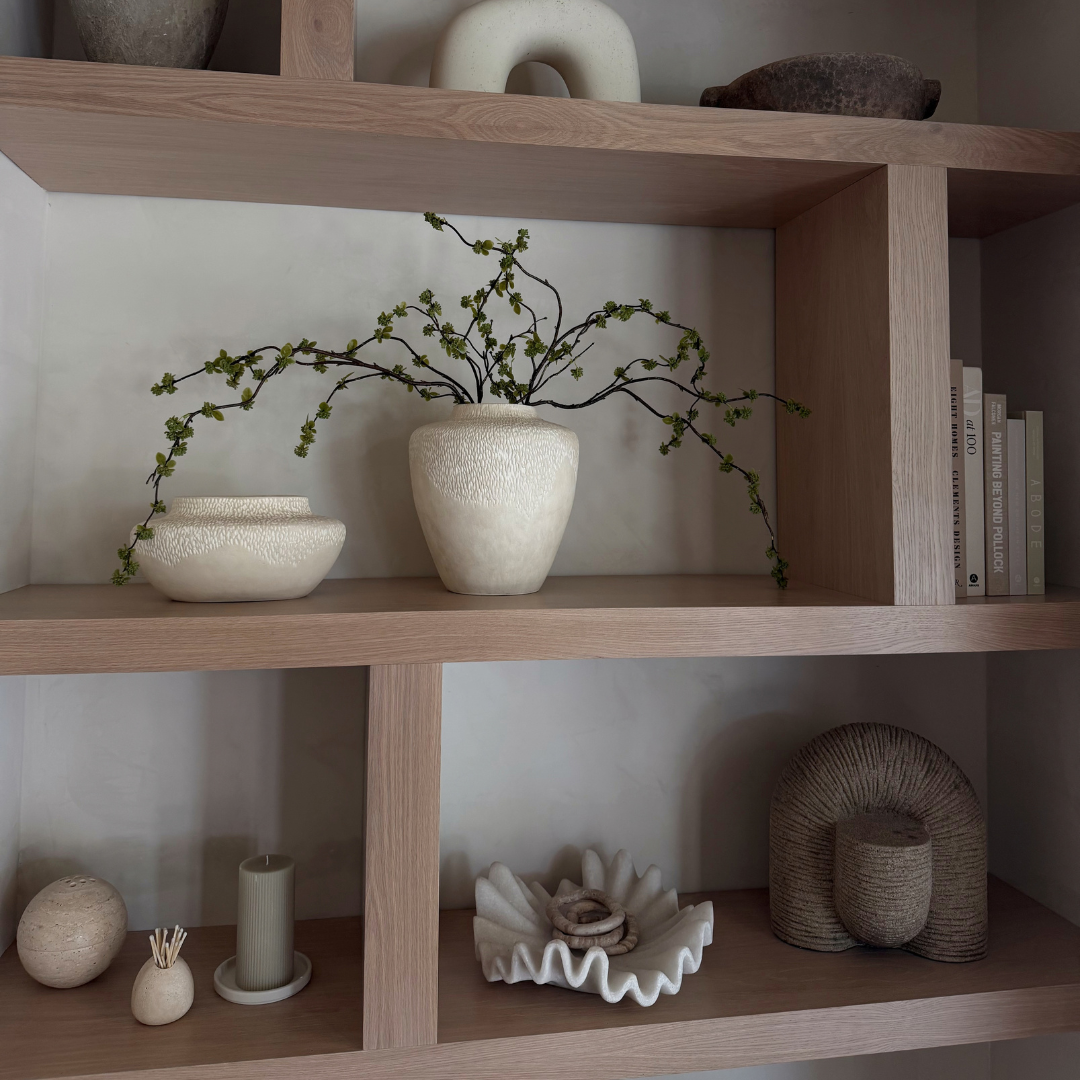 A light wood open shelving unit styled with terracotta neutral vases, a small bowl, books, and a dried green branch arrangement in the center vase.