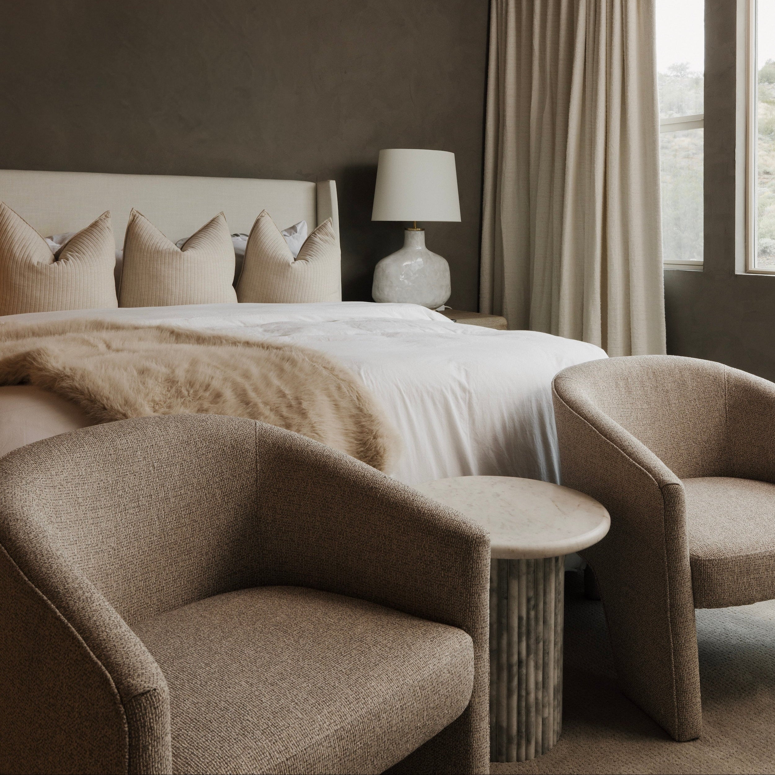 Elegant Reeve Linen Bed with upholstered headboard, white bedding, faux fur throw, and two textured armchairs.