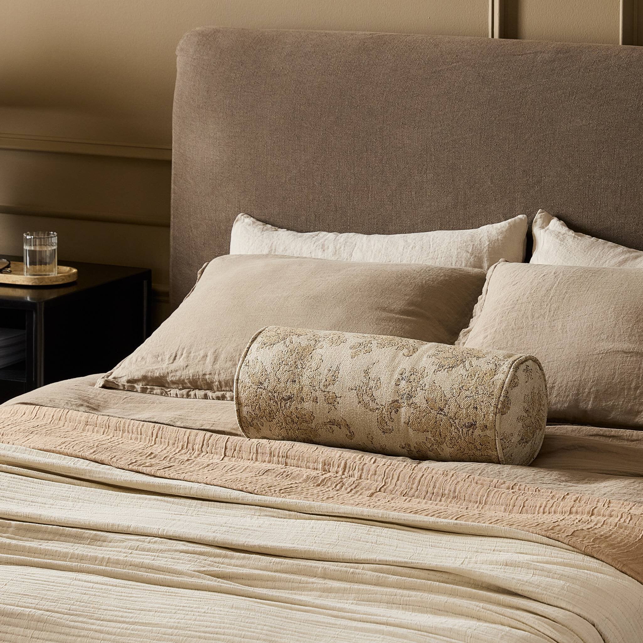 Neatly made bed with decorative pillows and a cylindrical bolster on a neutral-toned headboard.