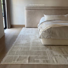 A light beige area rug with a subtle geometric pattern and fringed edges, styled in a modern bedroom next to a bed with neutral-toned bedding.