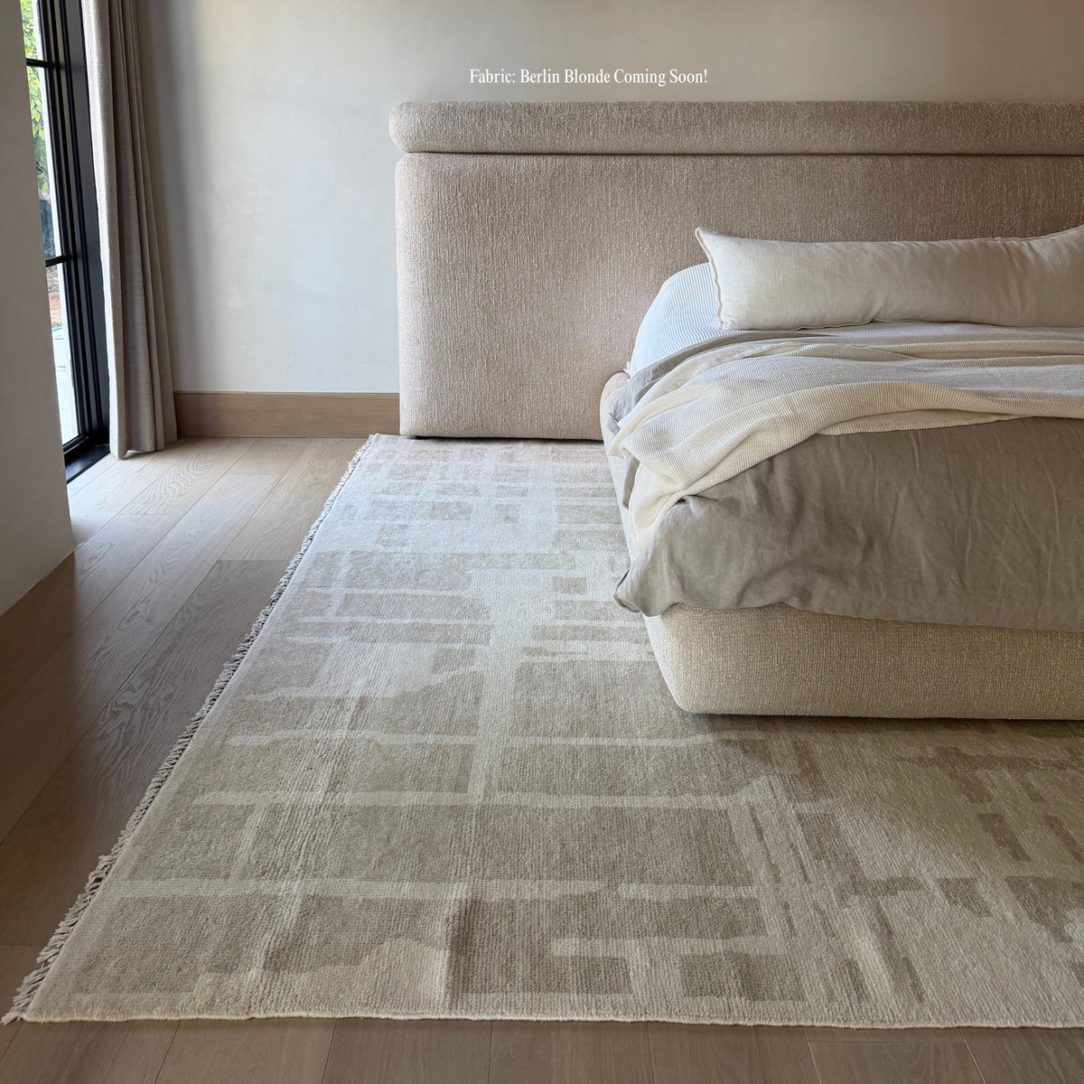 A light beige area rug with a subtle geometric pattern and fringed edges, styled in a modern bedroom next to a bed with neutral-toned bedding.