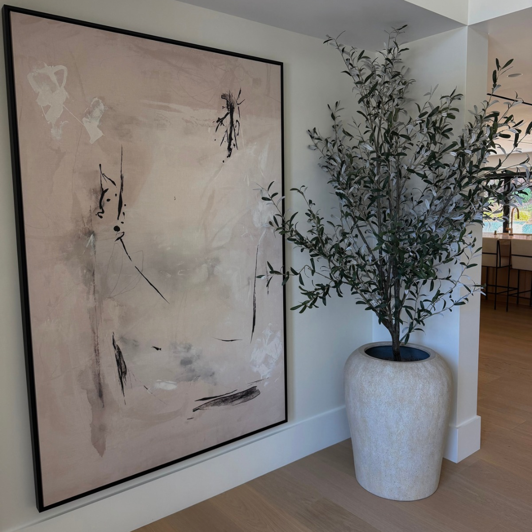 A large framed abstract wall art piece with soft blush, beige, and gray tones and black brushstrokes, displayed in a bright interior next to a tall potted olive tree.
