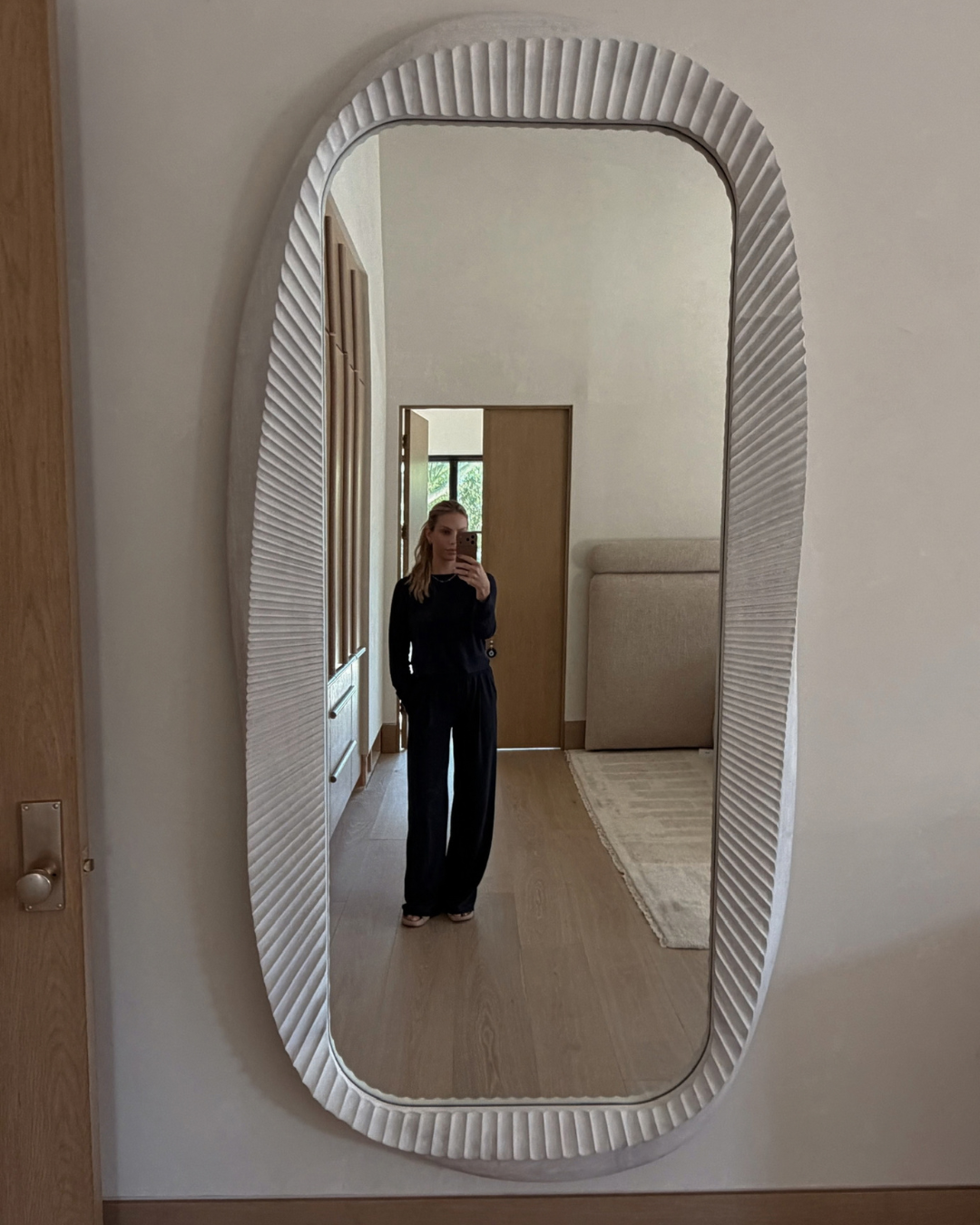 A large, vertically oriented oval wall mirror with a textured, ribbed white frame, shown in a home setting reflecting a person taking a mirror selfie.