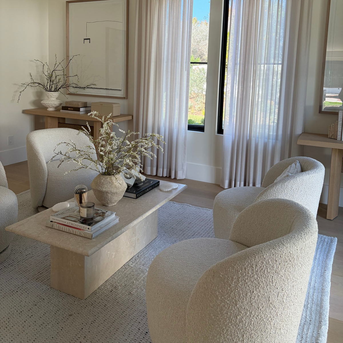 A modern living room featuring three rounded boucle armchairs in cream, a low concrete coffee table with books and a vase of dried branches, a light rug, sheer curtains, and a large framed abstract print on the wall.