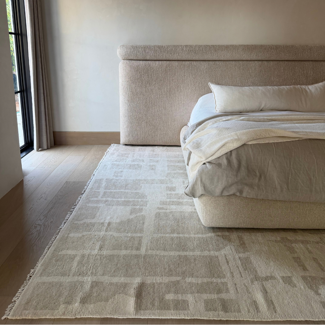 A large beige and off-white patterned area rug in a modern bedroom setting, styled under a matching upholstered bed with neutral bedding.