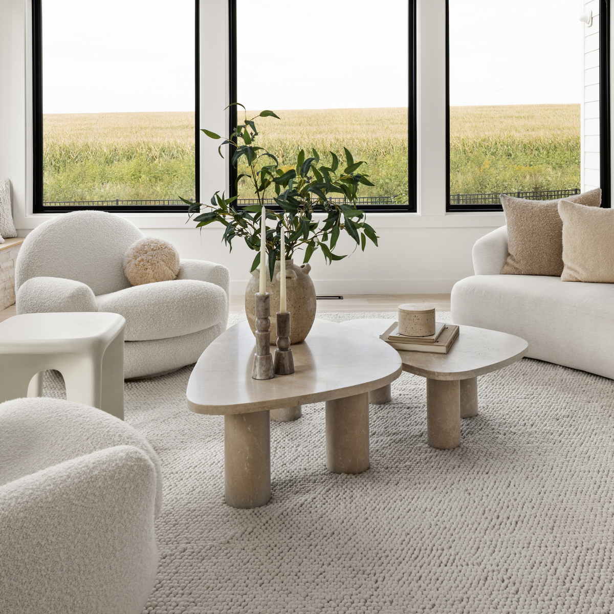 A modern living room with white textured furniture, including a curved armchair, a sofa, and a low organic-shaped coffee table, set in front of large windows overlooking a field.