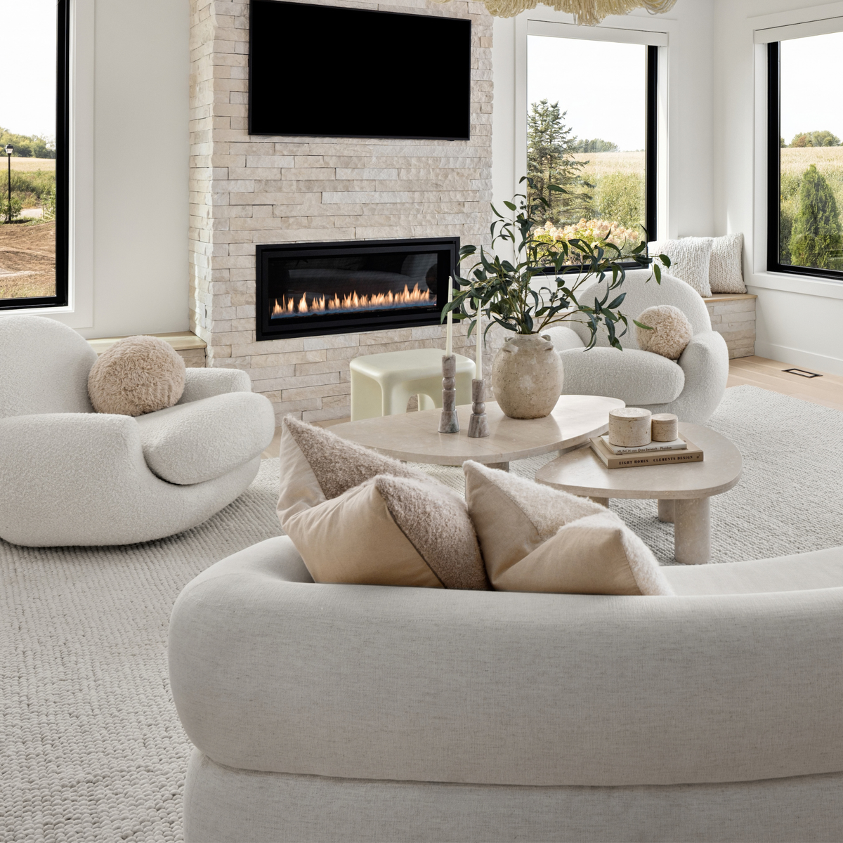 A bright, modern living room featuring a light beige upholstered curved sofa in the foreground, two matching rounded armchairs, a round wooden coffee table with a vase of greenery, and a stone fireplace with a built-in linear firebox and mounted TV.