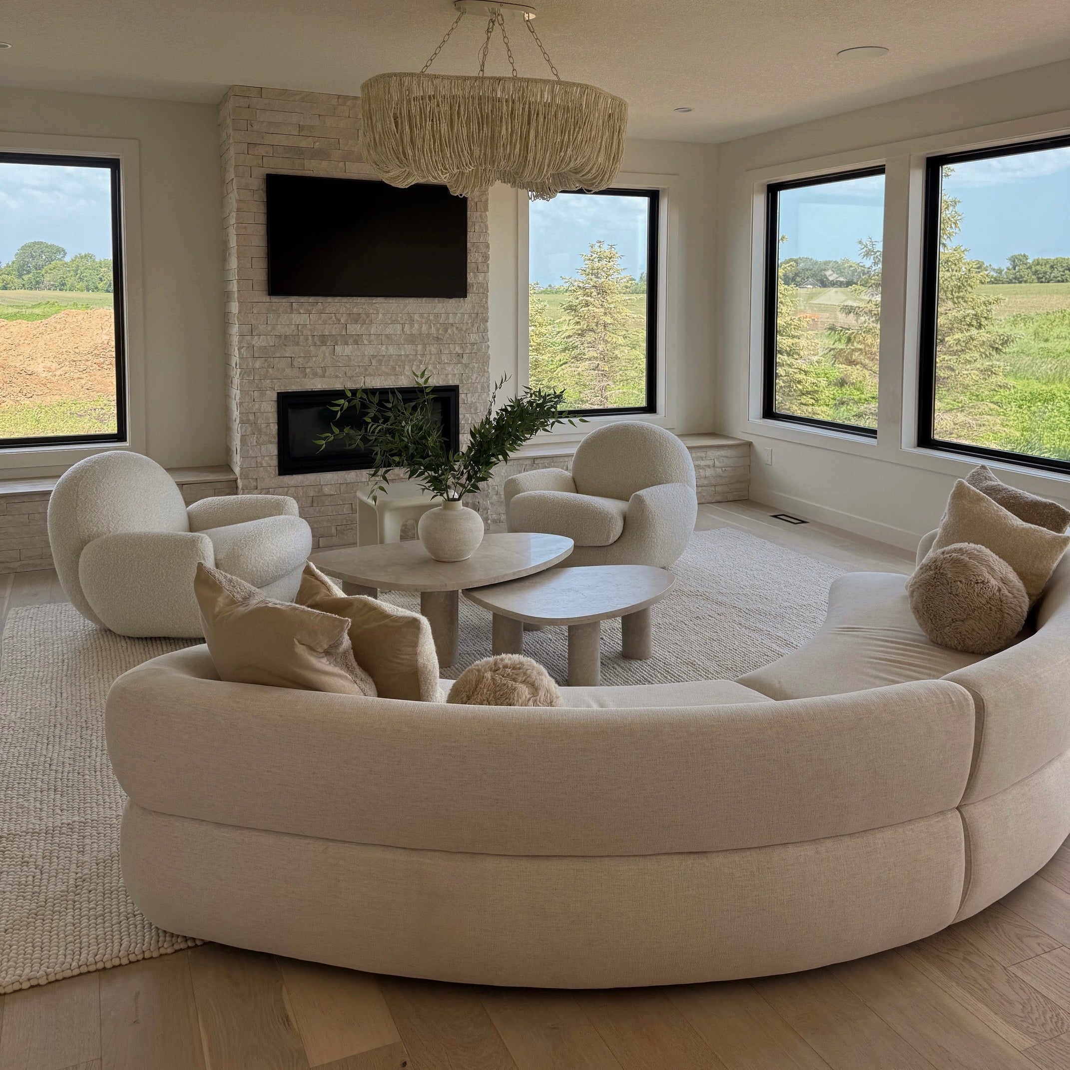 A large curved upholstered sofa in a light cream or off-white fabric is shown in a bright modern living room with large windows, a stone fireplace, round accent chairs, and a chandelier. No people or animals are present.