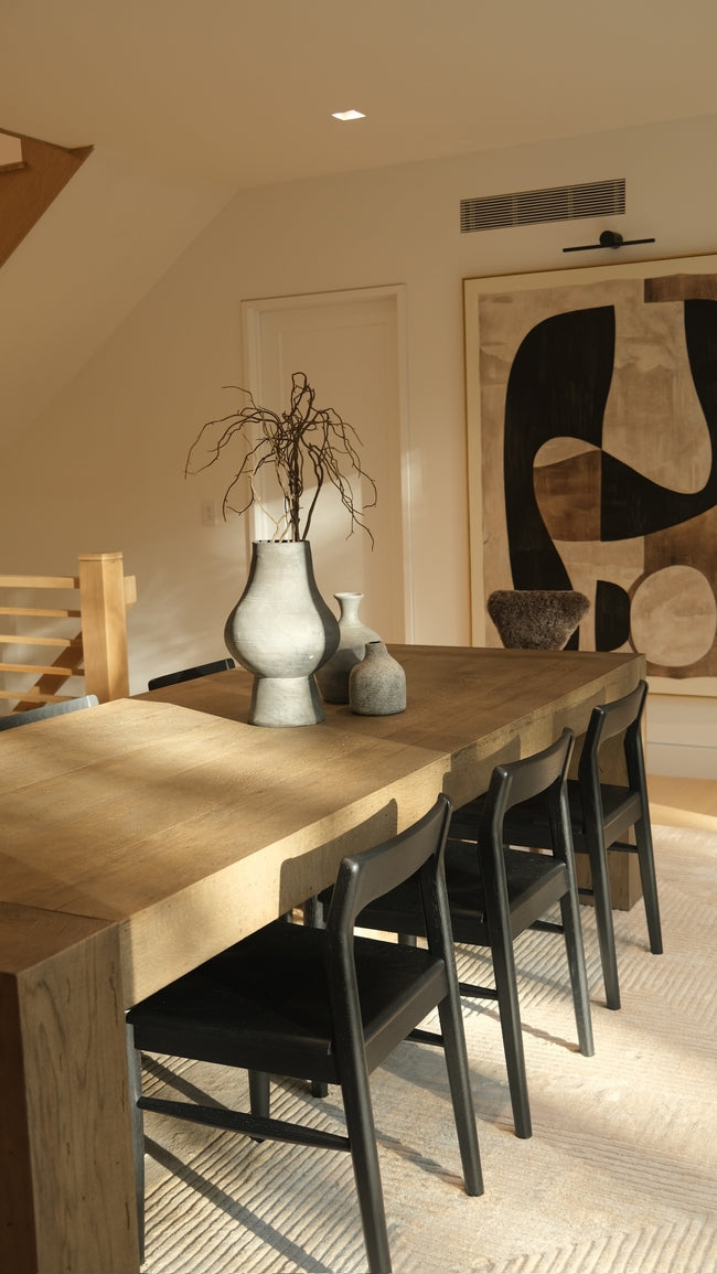 Dining area with a long wooden table and black chairs in a room with abstract art on the wall.