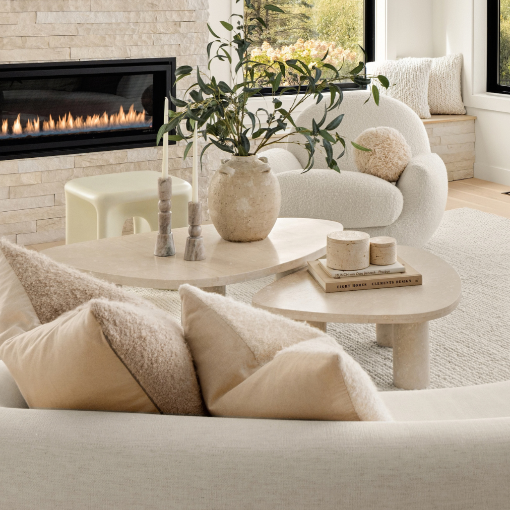 A bright, modern living room featuring a large cream boucle sofa in the foreground, two matching curved armchairs, a round coffee table with neutral pillows, a light stone fireplace with a built-in fire, and large windows looking out onto green fields.