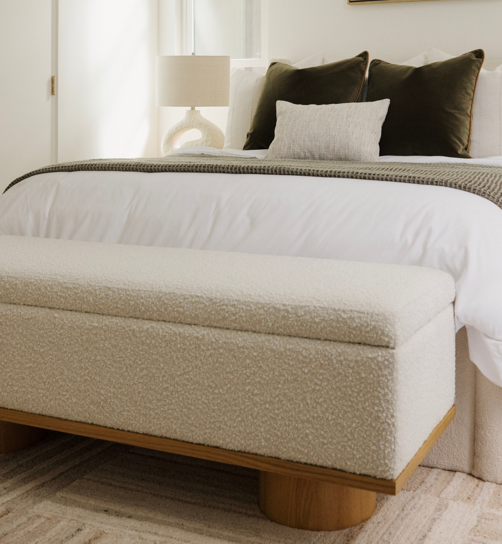 Beige upholstered bench at the foot of a bed with white bedding and brown pillows.
