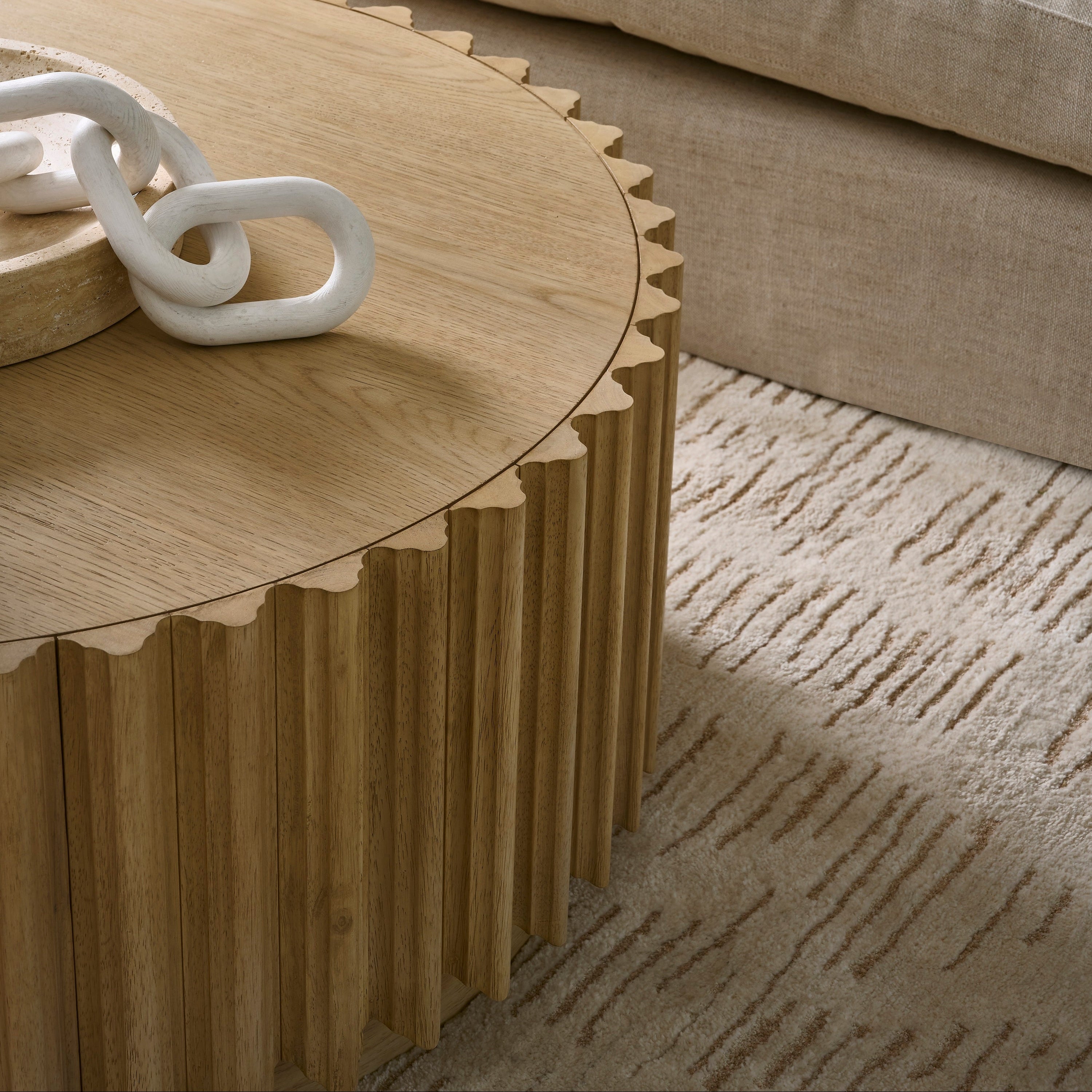 A close-up of a round wooden coffee table with a fluted slatted base and scalloped edge, styled in a modern living room next to a beige sofa and textured rug.
