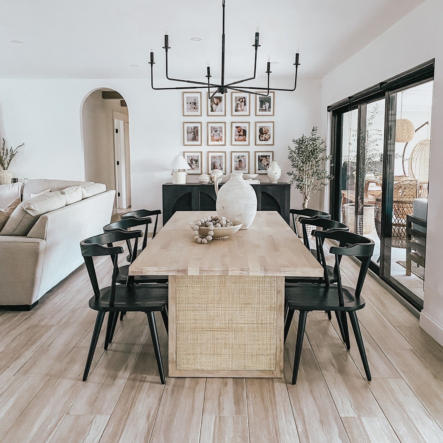 A modern dining room featuring a rectangular light wood table with a woven rattan base, surrounded by black chairs under a black multi-arm chandelier. The room has light wood floors, white walls, and large sliding glass doors.