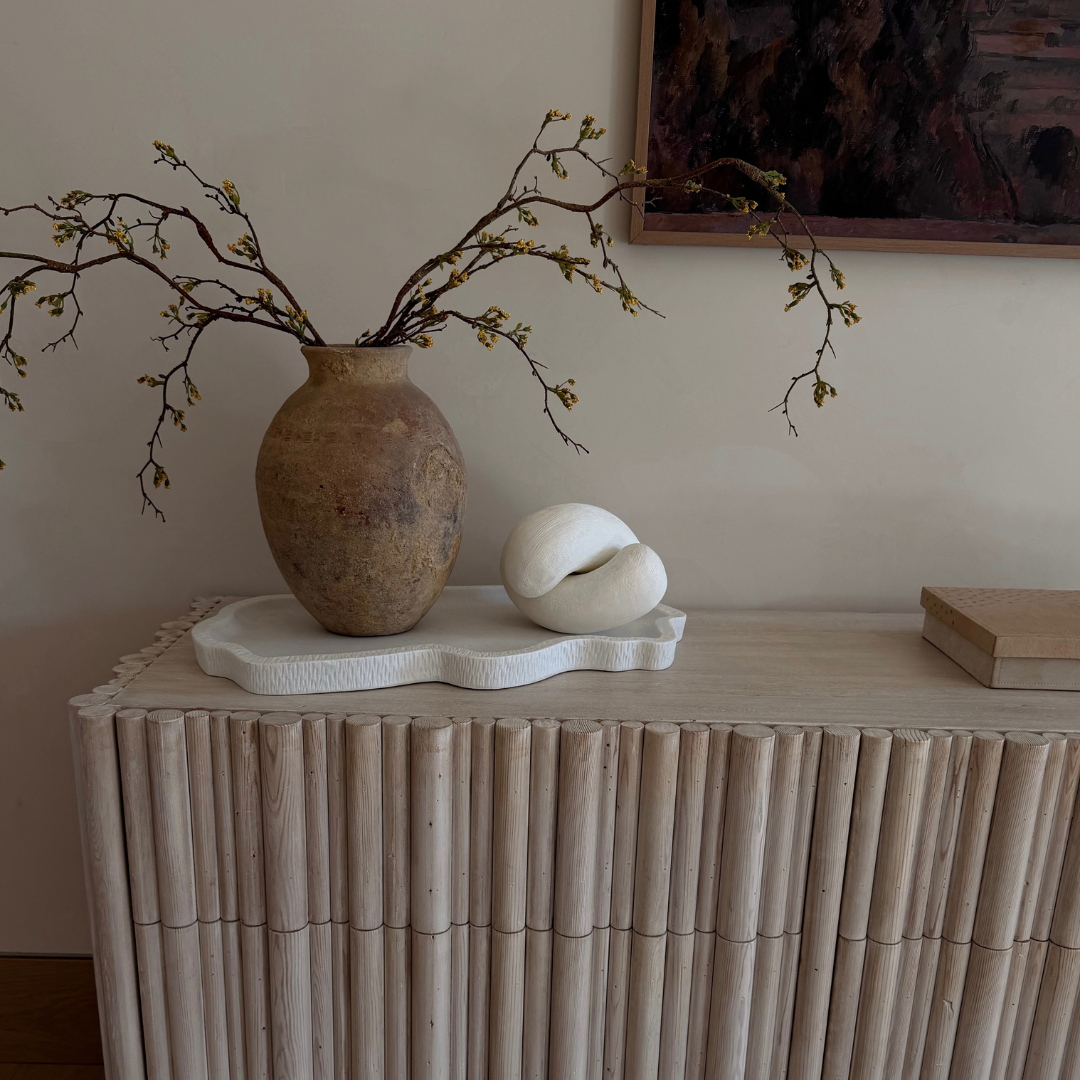 A light natural wood reeded dresser with a slatted front, styled with a textured brown ceramic vase holding bare branches, a white sculptural object, and a small book against a beige wall.