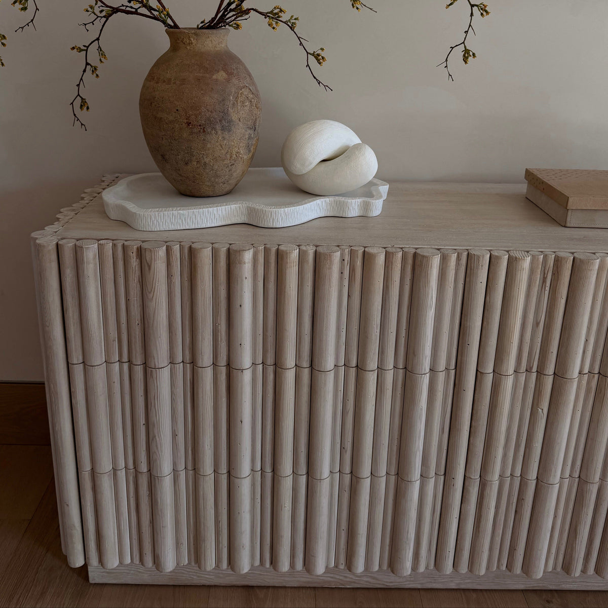 A light natural wood dresser with a vertically fluted bamboo-like front, styled with a brown ceramic vase, white sculptural decor, and branches in a neutral interior setting.