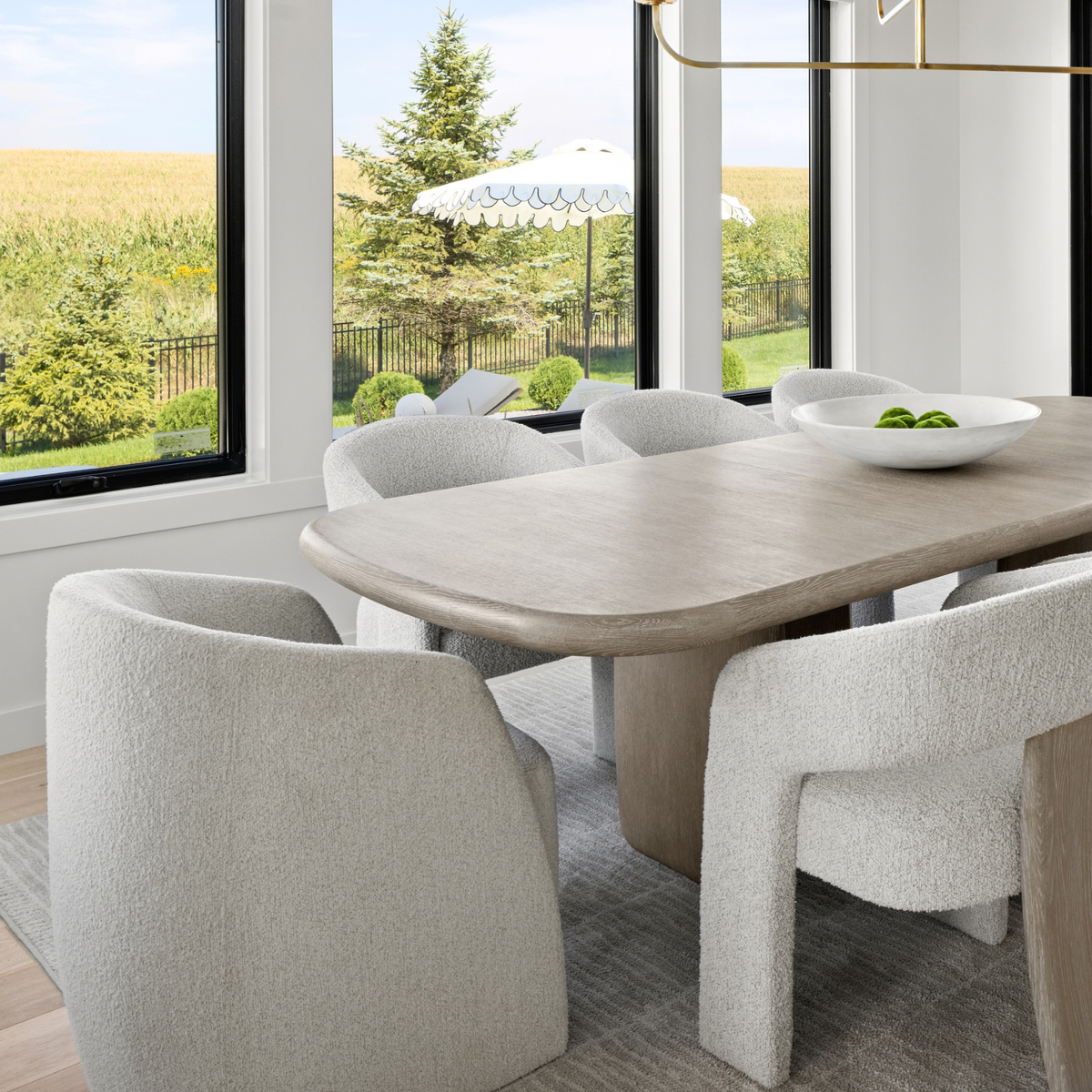 A modern dining room scene with a light wood oval table surrounded by textured off-white boucle chairs, set in a bright room with large windows overlooking a yard.