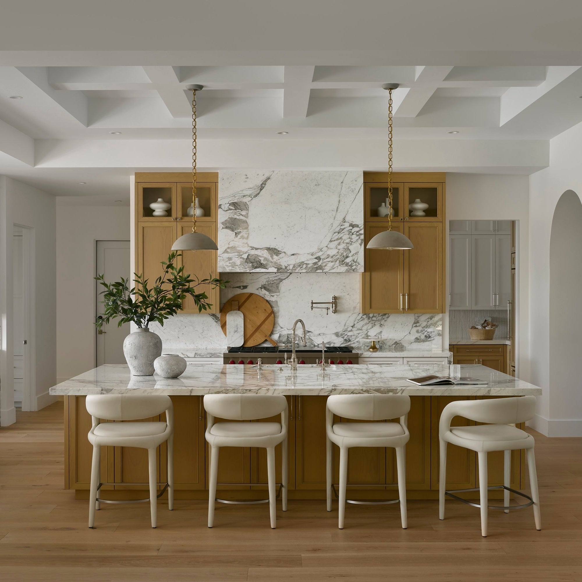 A modern kitchen island with a white marble countertop and backsplash, surrounded by four white bar stools with light wood frames. The kitchen features light wood cabinetry, a white coffered ceiling, and light wood flooring.