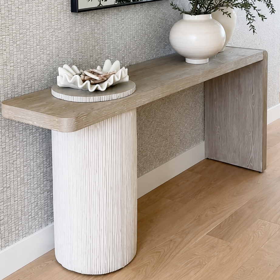 A modern console table with a light wood top and a textured white cylindrical pedestal base, styled in a home entryway with a black-framed mirror, white ceramic vase, and decorative bowl.