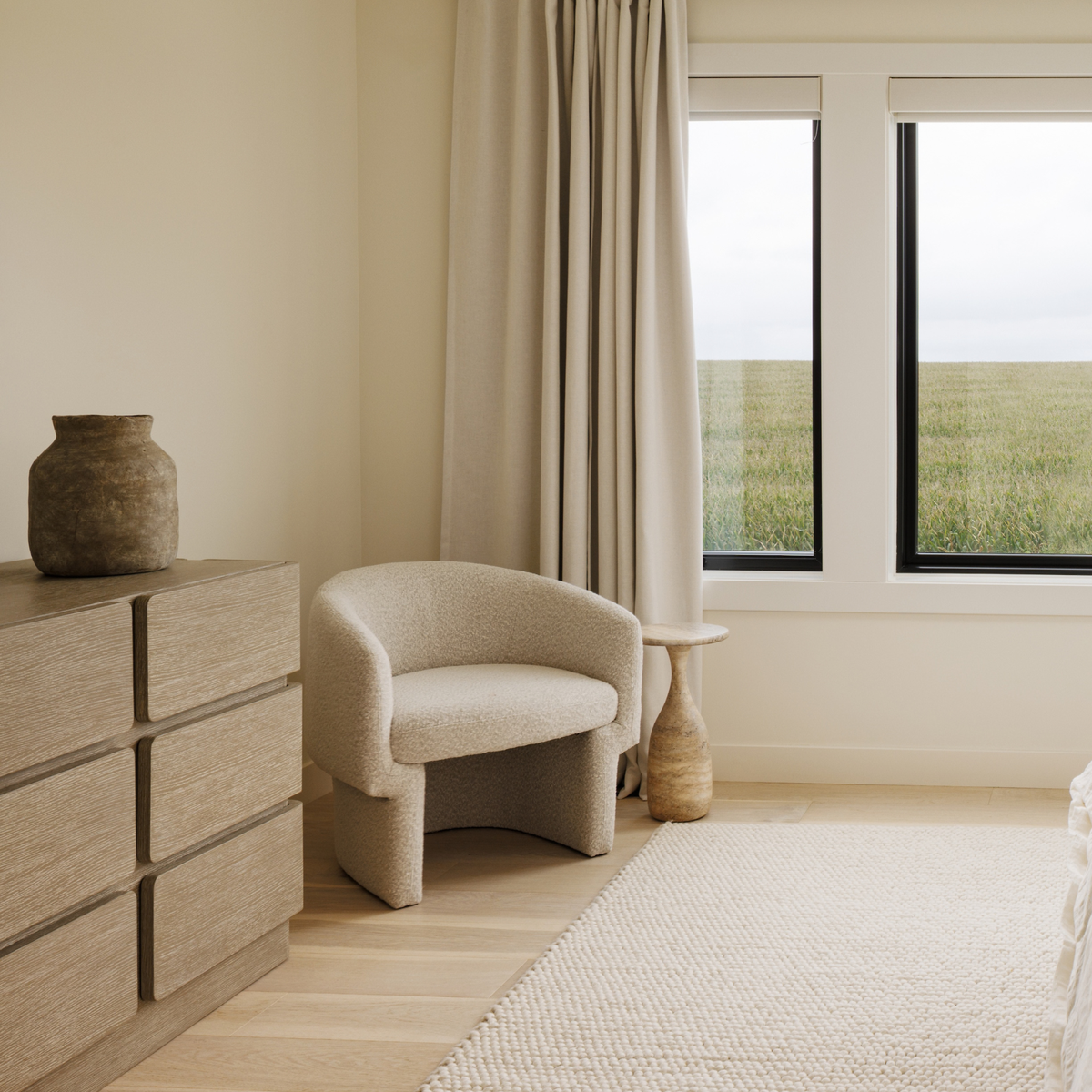 A modern bedroom corner featuring a curved upholstered armchair in a textured beige fabric, positioned next to a light wood dresser and a small woven side table. Large windows with beige drapes look out onto a grassy field, and a neutral rug covers the light wood floor.