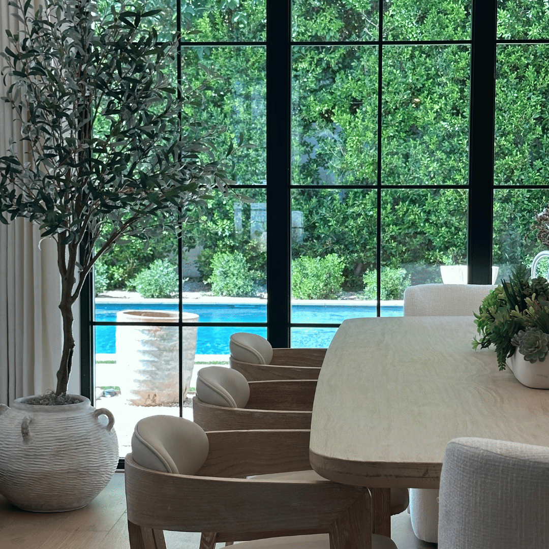Dining area with wooden table and chairs, potted plants, and large windows overlooking greenery.