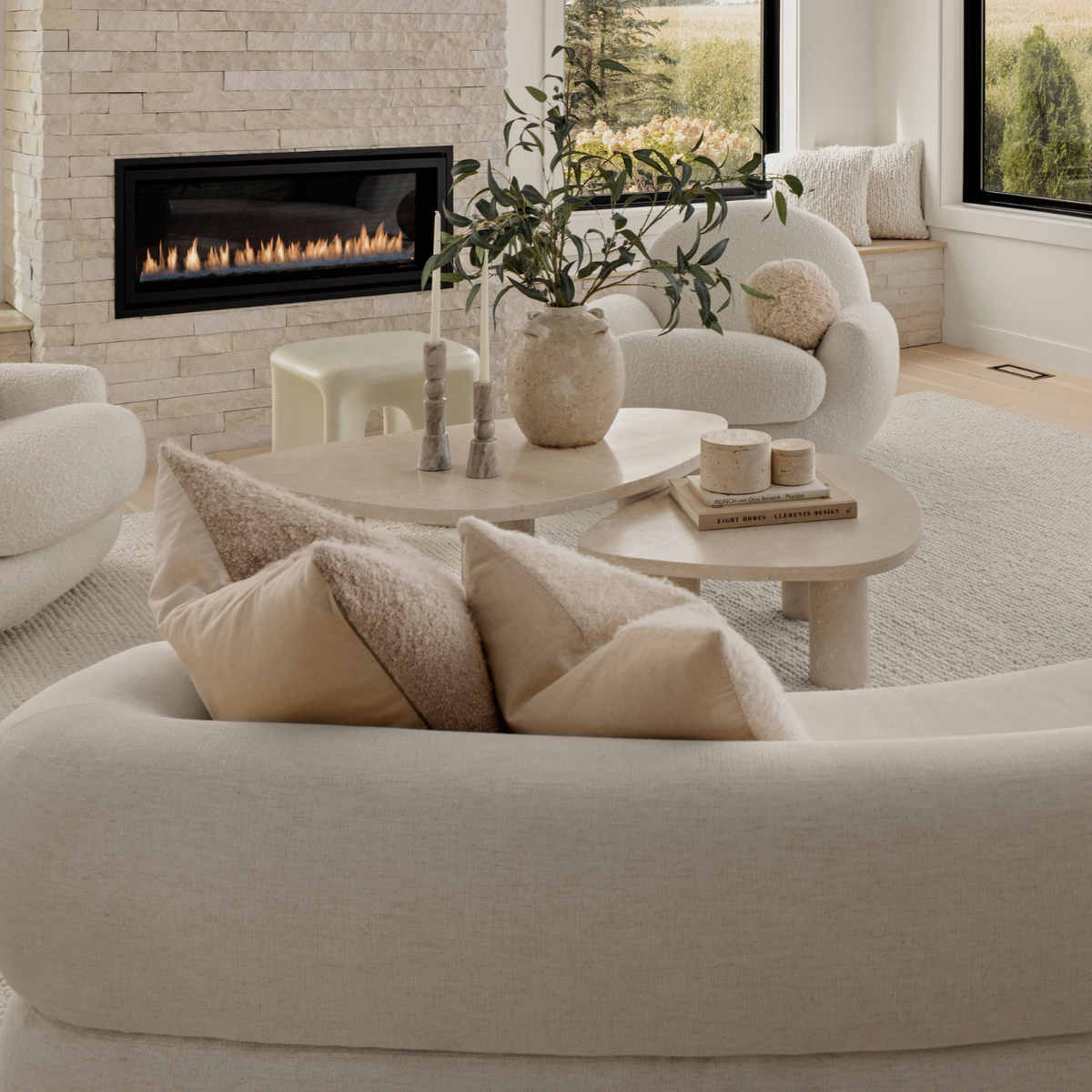 A bright, modern living room featuring a curved cream sofa in the foreground, a matching round coffee table with neutral-toned decor, white upholstered armchairs, a stone fireplace with a mounted TV, and large windows overlooking a green field.