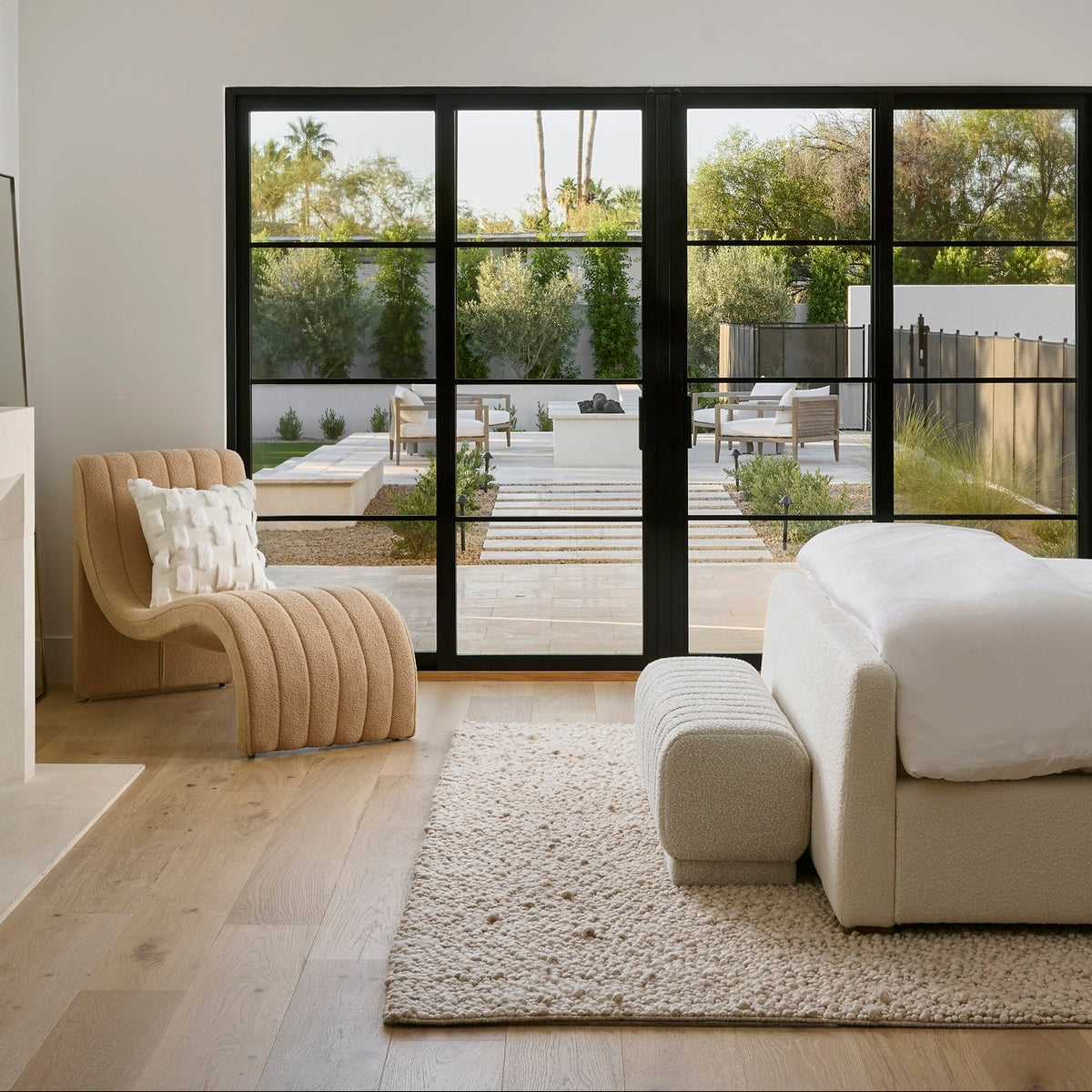 A modern bedroom interior with a cream upholstered bed, matching pillows, a textured beige rug, a striped armchair, a fireplace, and large black-framed glass doors looking out onto a patio and trees.